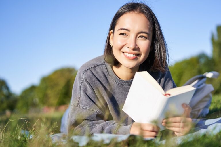 Portrait Of Cute Korean Girl, Reading In Park While Lying On Grass, Relaxing With Favorite Book In Hands, Smiling Happily